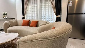A modern living room with a beige sectional sofa, a small round table, and a large window with sheer curtains. - LivLuxe Apartments 5 (Kampala)