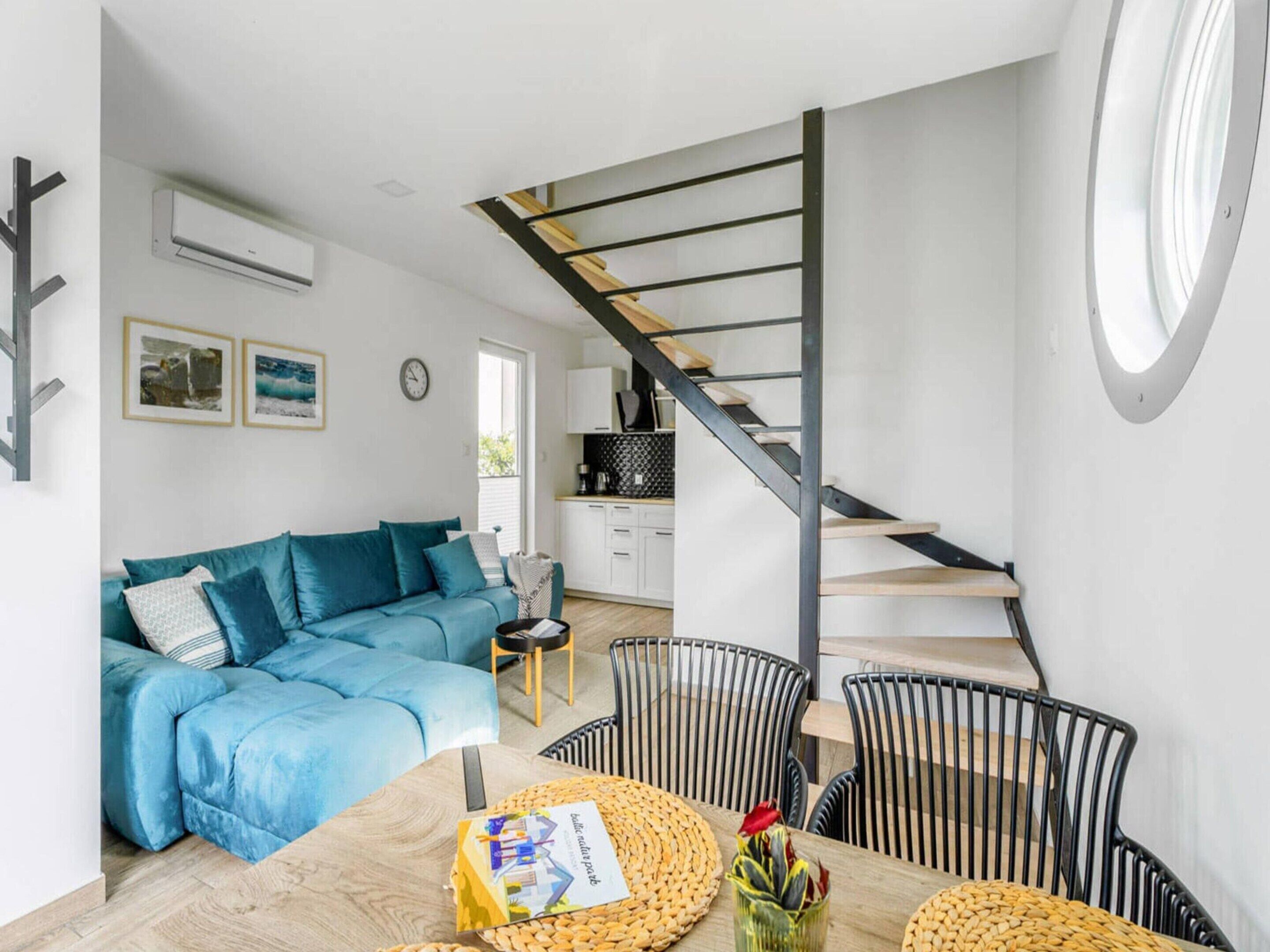Un espacio de estar moderno con un sofá azul, una mesa de comedor de madera con individuales tejidos, una escalera de metal negro y una zona de cocina al fondo.