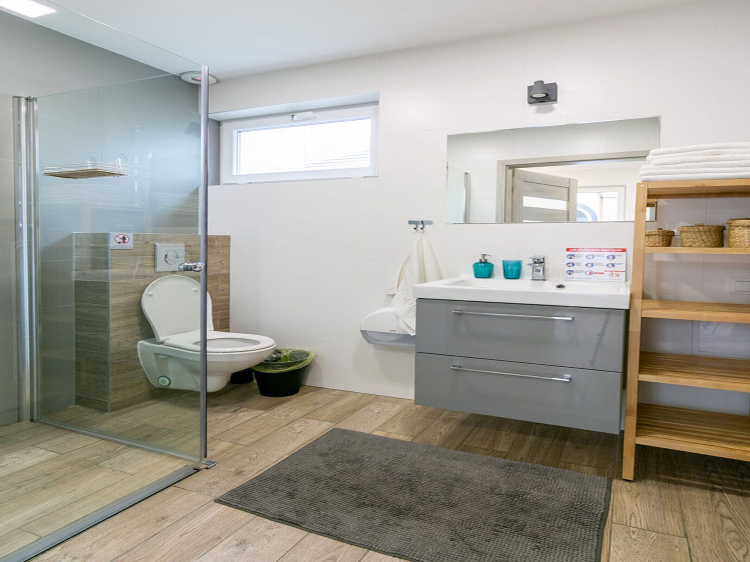 Ein modernes Badezimmer mit Duschkabine aus Glas, Toilette, Waschbecken mit Spiegel und Holzregal mit Körben.
