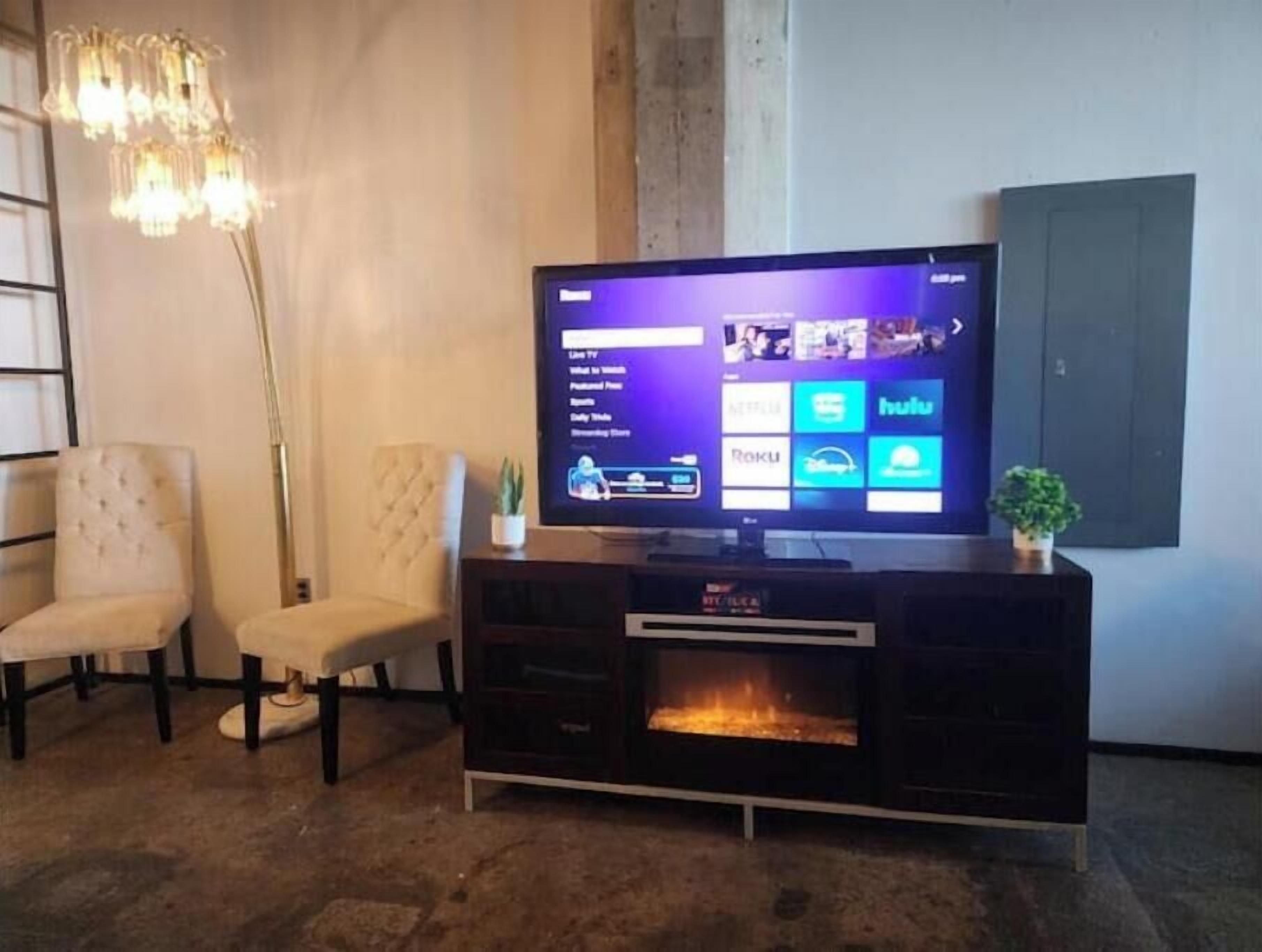 Una sala de estar con un televisor de pantalla plana sobre un soporte negro, una chimenea, dos sillones blancos y una lámpara de pie alta.
