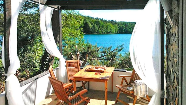 An outdoor patio with a wooden table and chairs, overlooking a lake with trees in the background. - A Wonderful Holiday Home With Lake Access (Wejherowo)