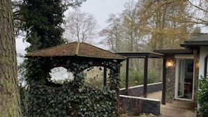 A covered outdoor area with a wooden structure partially covered in ivy, a stone pathway, and a building entrance with glass doors. - A Wonderful Holiday Home With Lake Access (Wejherowo)