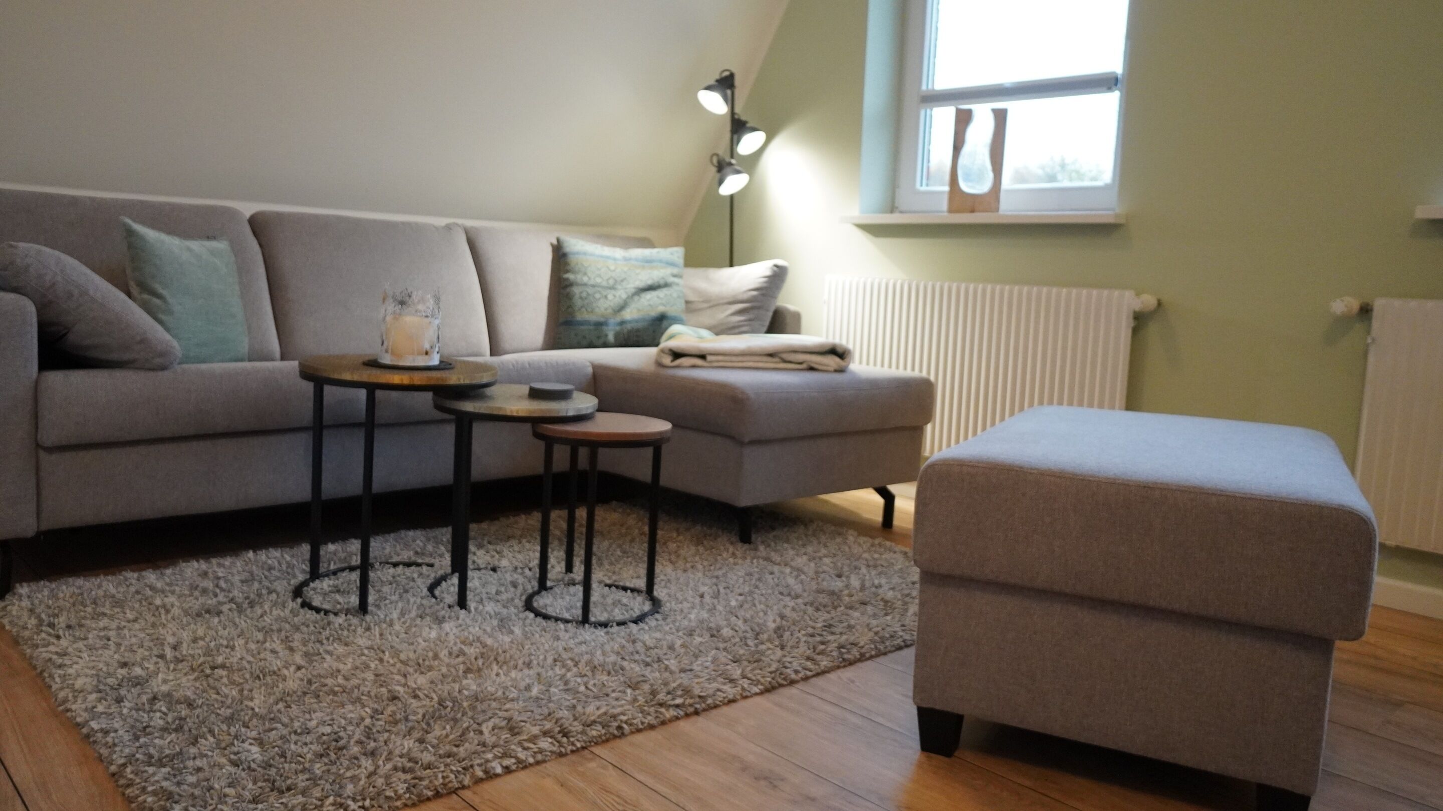 Un salon avec un canapé gris, une petite table ronde, un pouf gris et une fenêtre avec des stores.