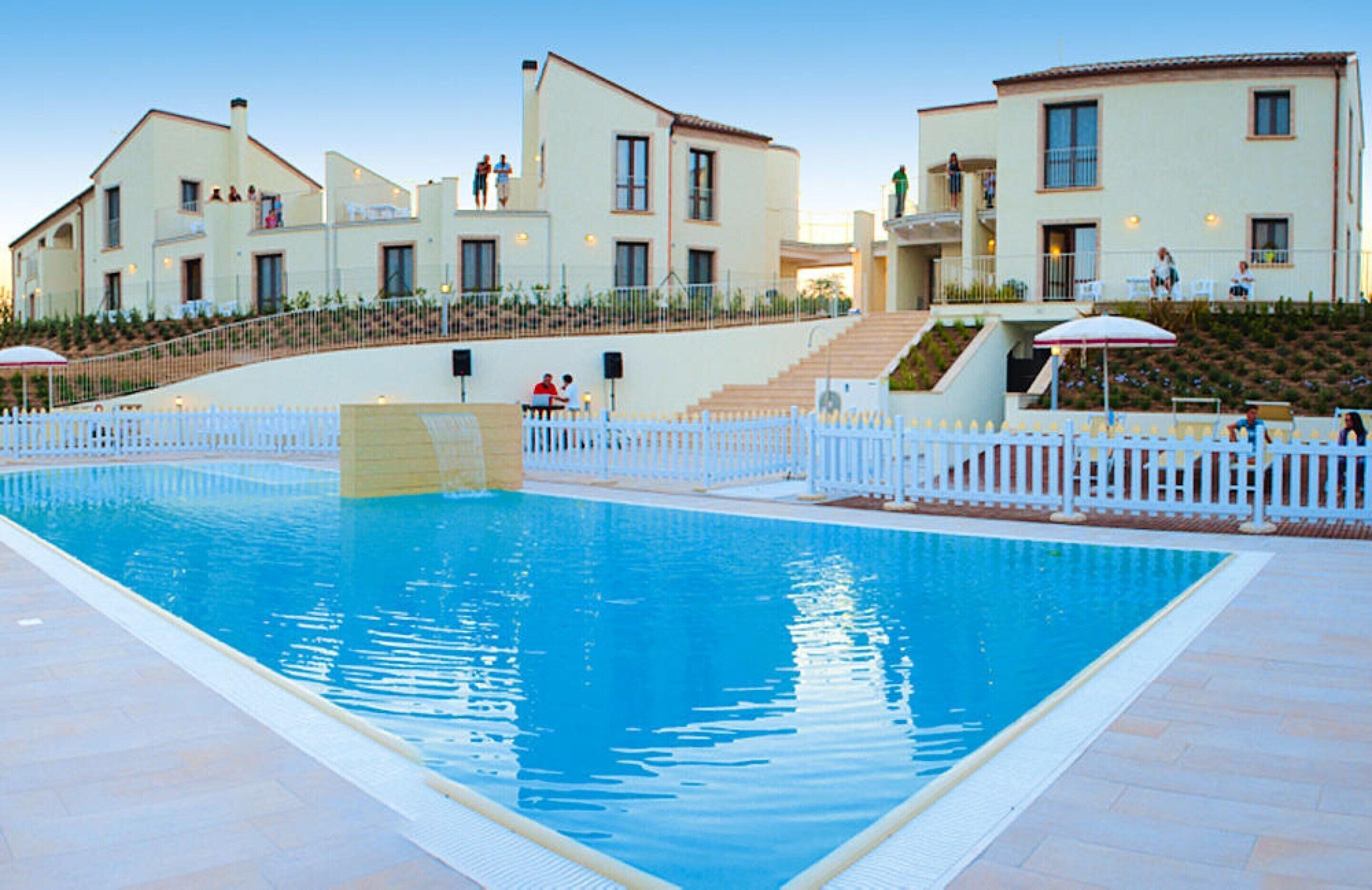 Une piscine entourée d’une clôture blanche, avec des marches menant à un bâtiment doté de plusieurs balcons.