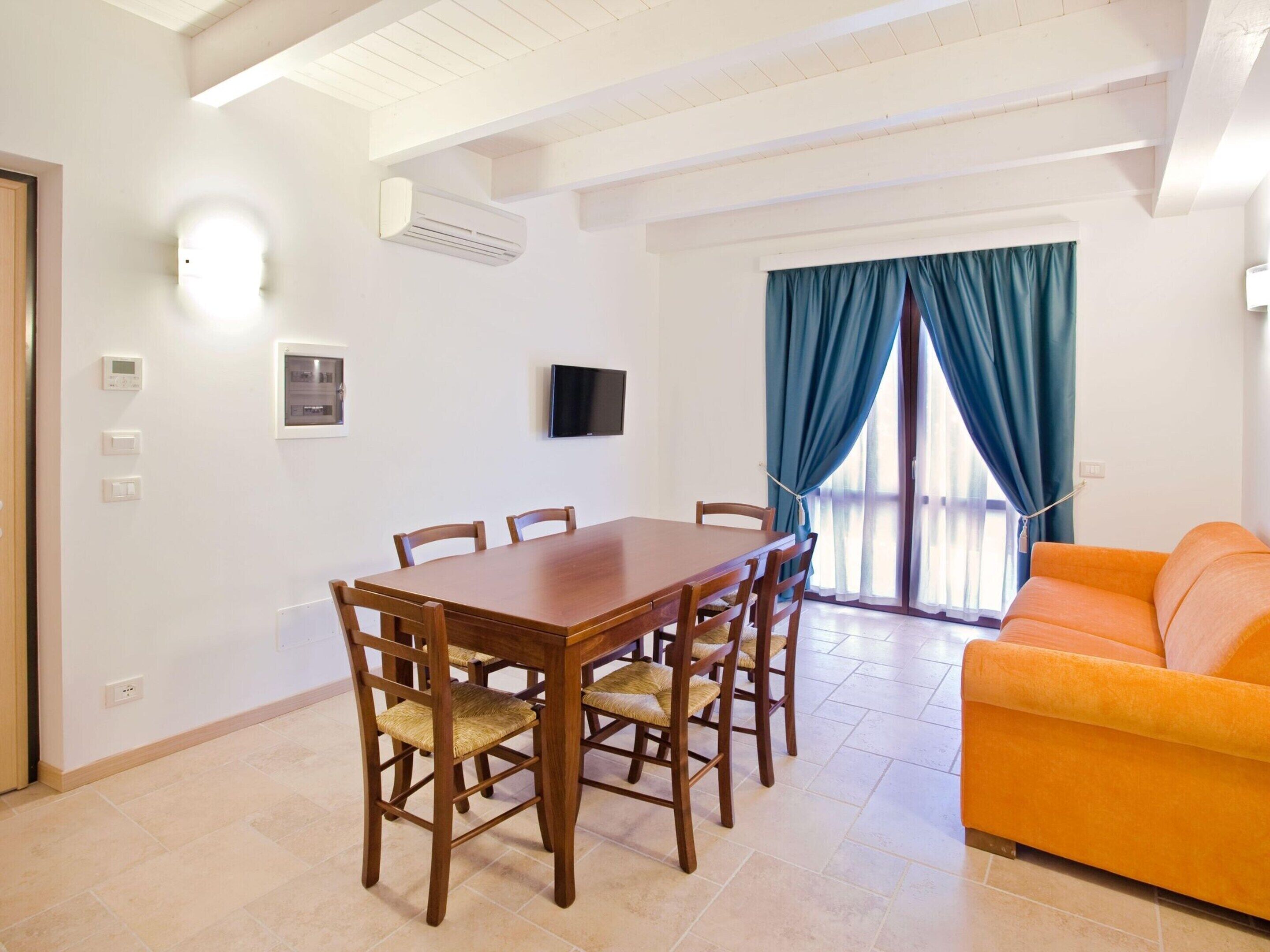 A dining area with a wooden table and chairs, a television mounted on the wall, and a sofa in the adjacent living area.