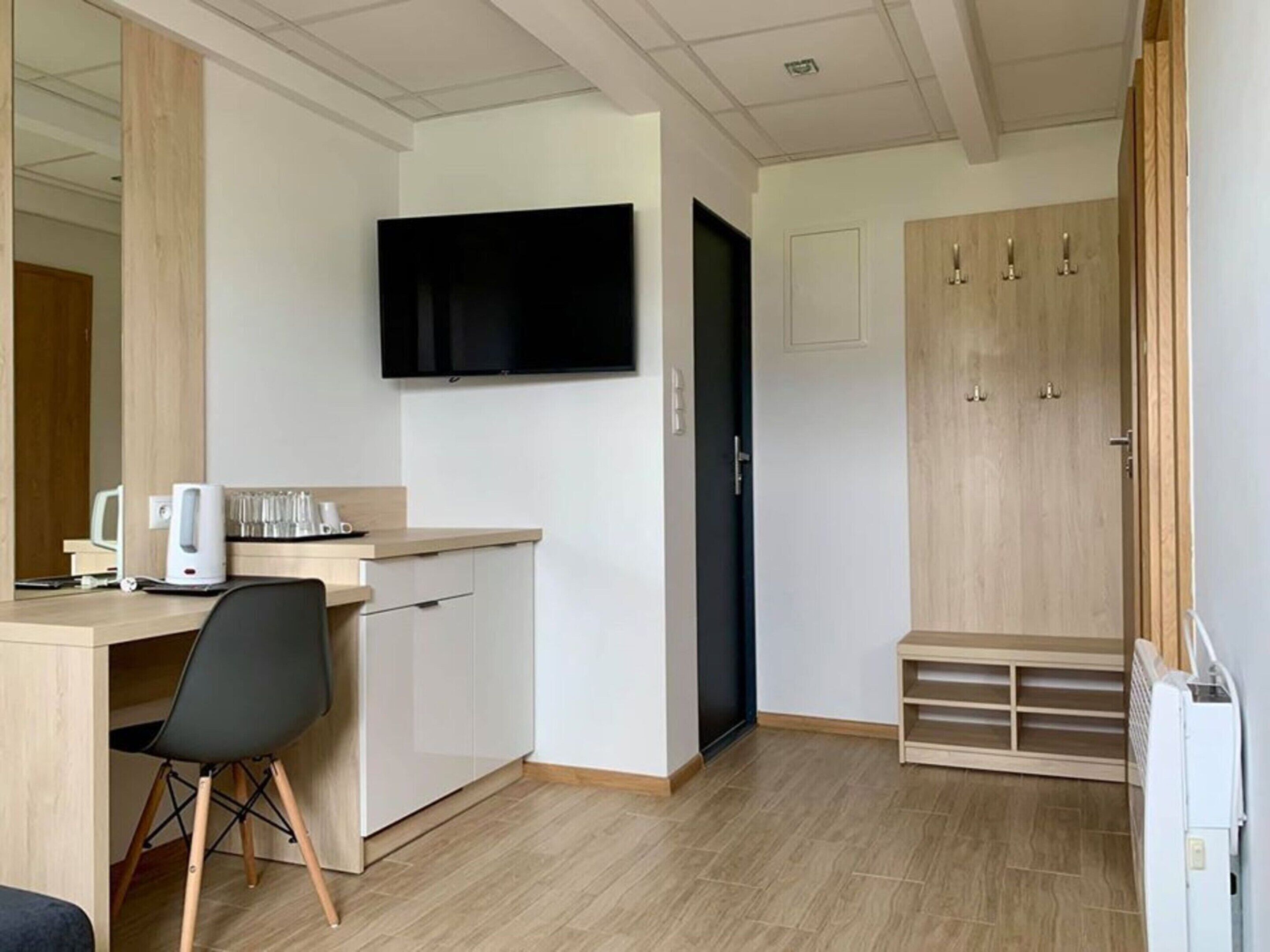 A modern hotel room with a flat-screen TV mounted on the wall, a wooden desk with a chair, a small kitchenette, and a wardrobe with a mirror.