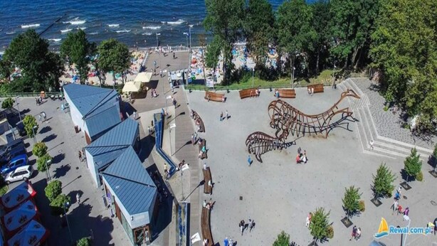 An aerial view of a coastal promenade with a large metal sculpture, a parking lot, and a view of the sea.