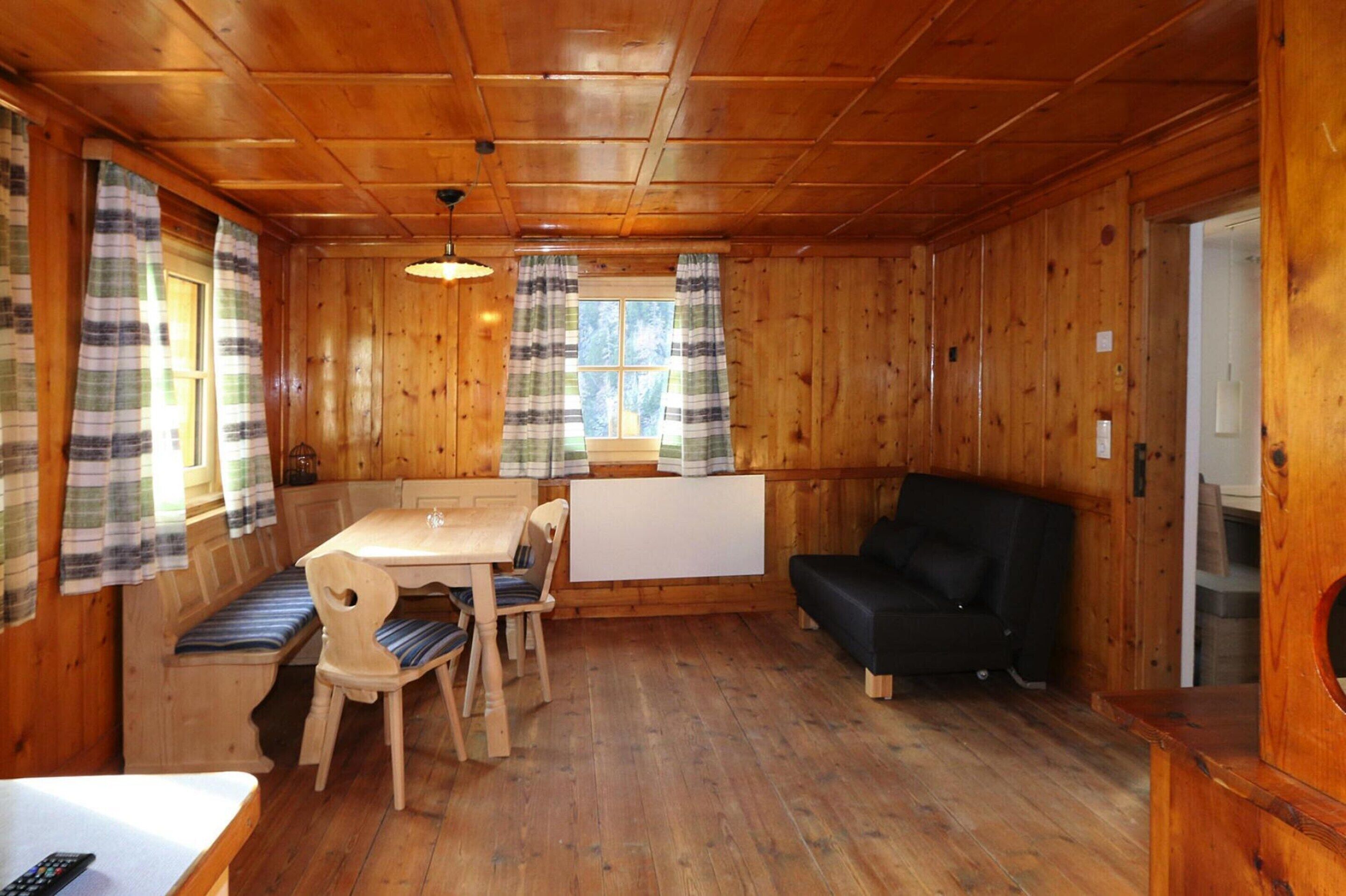 A wooden cabin interior with a dining table, chairs, a black sofa, and a window with curtains.