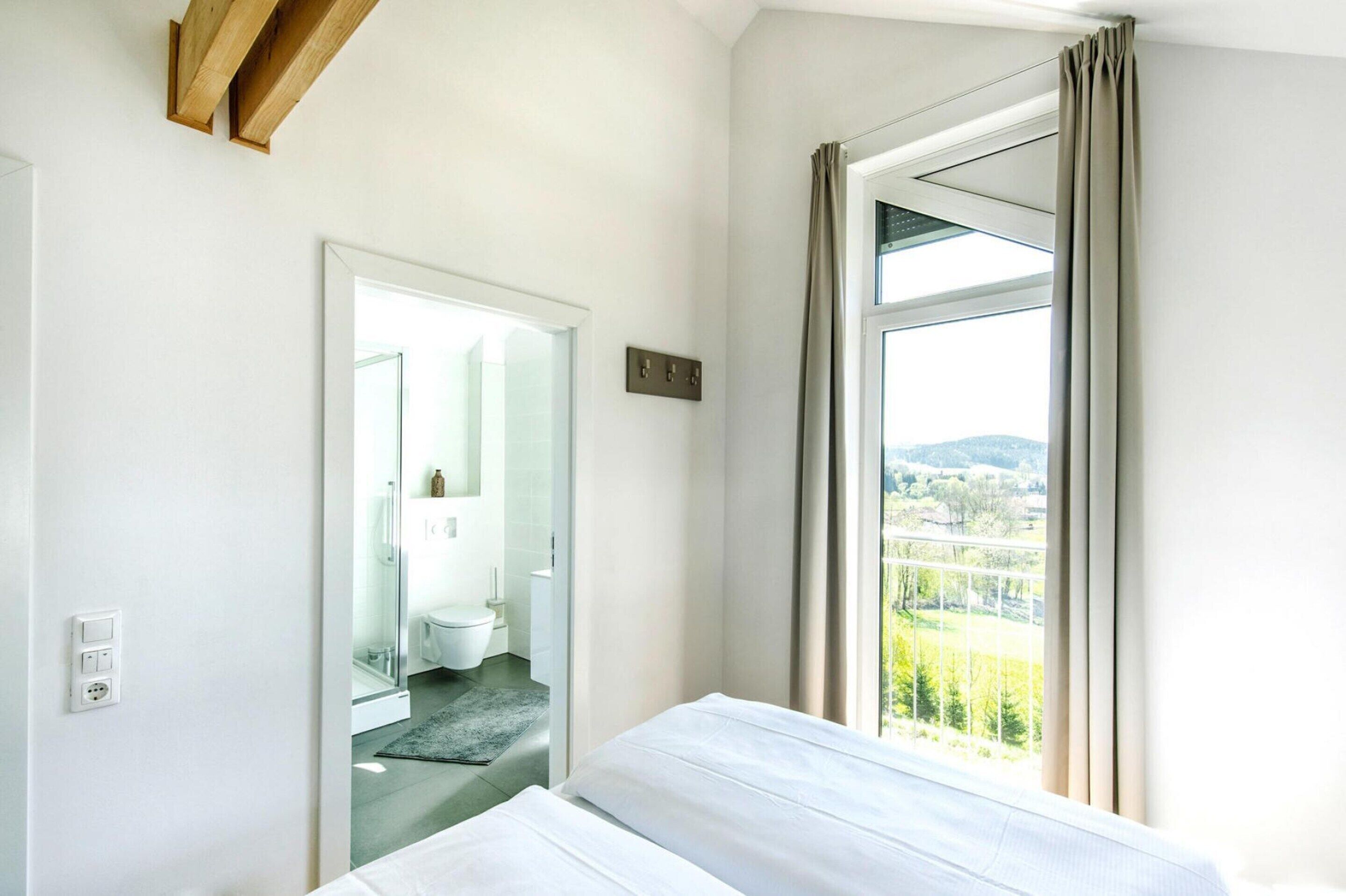 A hotel room with a bed, a window with curtains, a bathroom visible through an open door, and a view of a landscape.