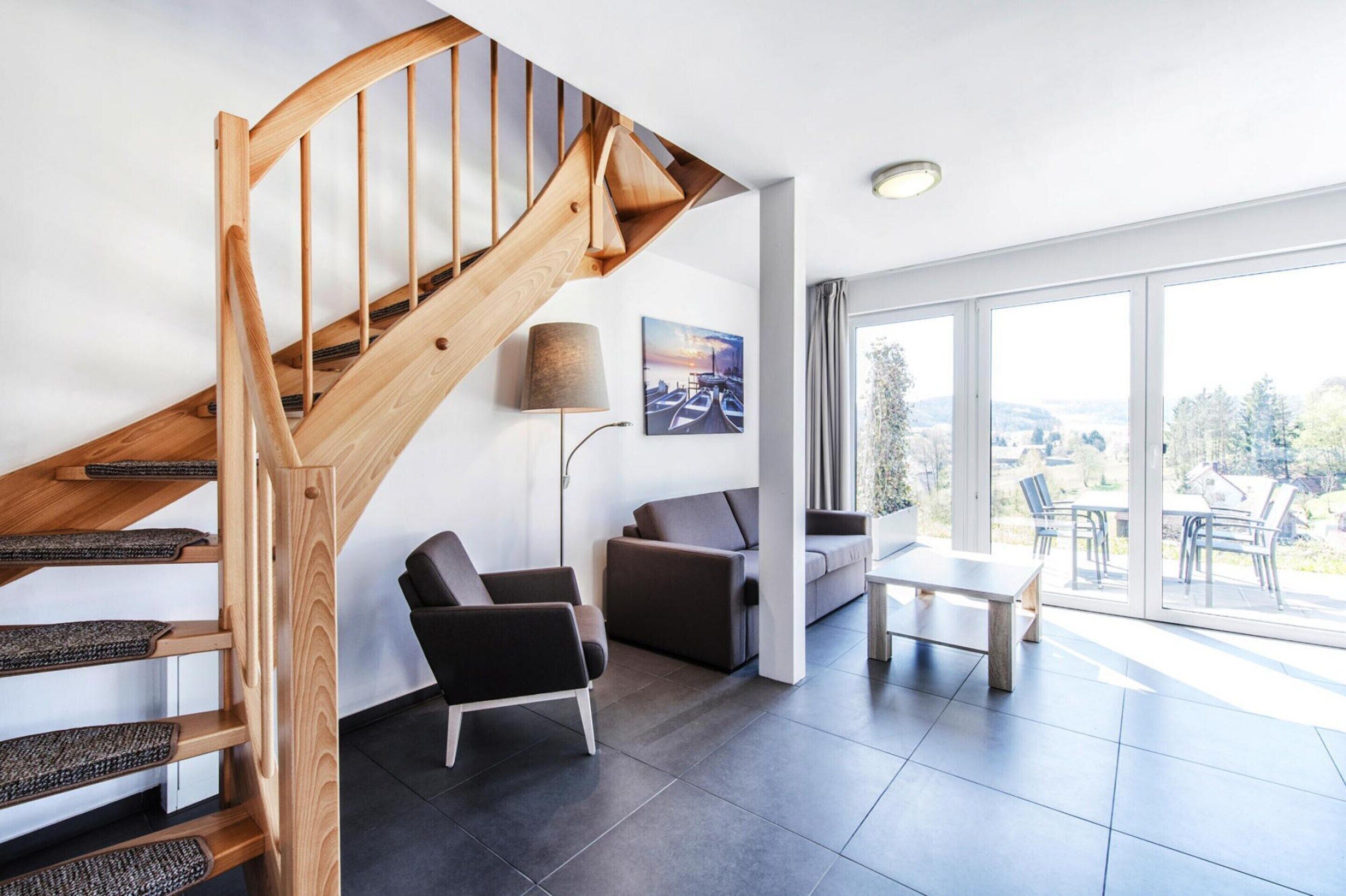 A modern living room with a wooden staircase, a sofa, a coffee table, and a large window offering a view of the outdoors.