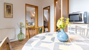A modern living room with a round table, a blue vase with flowers, a microwave, and a dining area with wooden chairs and a green vase.