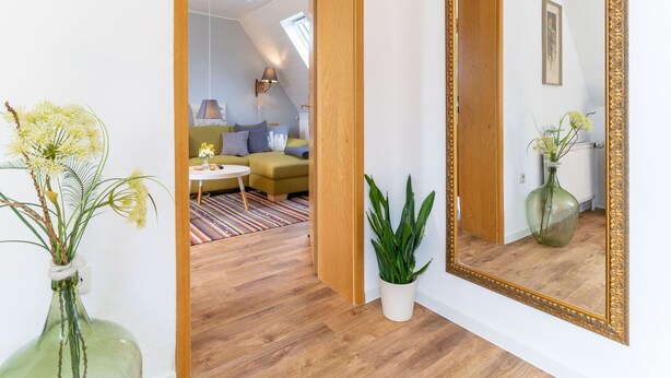 A room with a wooden floor, a green sofa, a white vase with flowers, and a large mirror with an ornate frame.