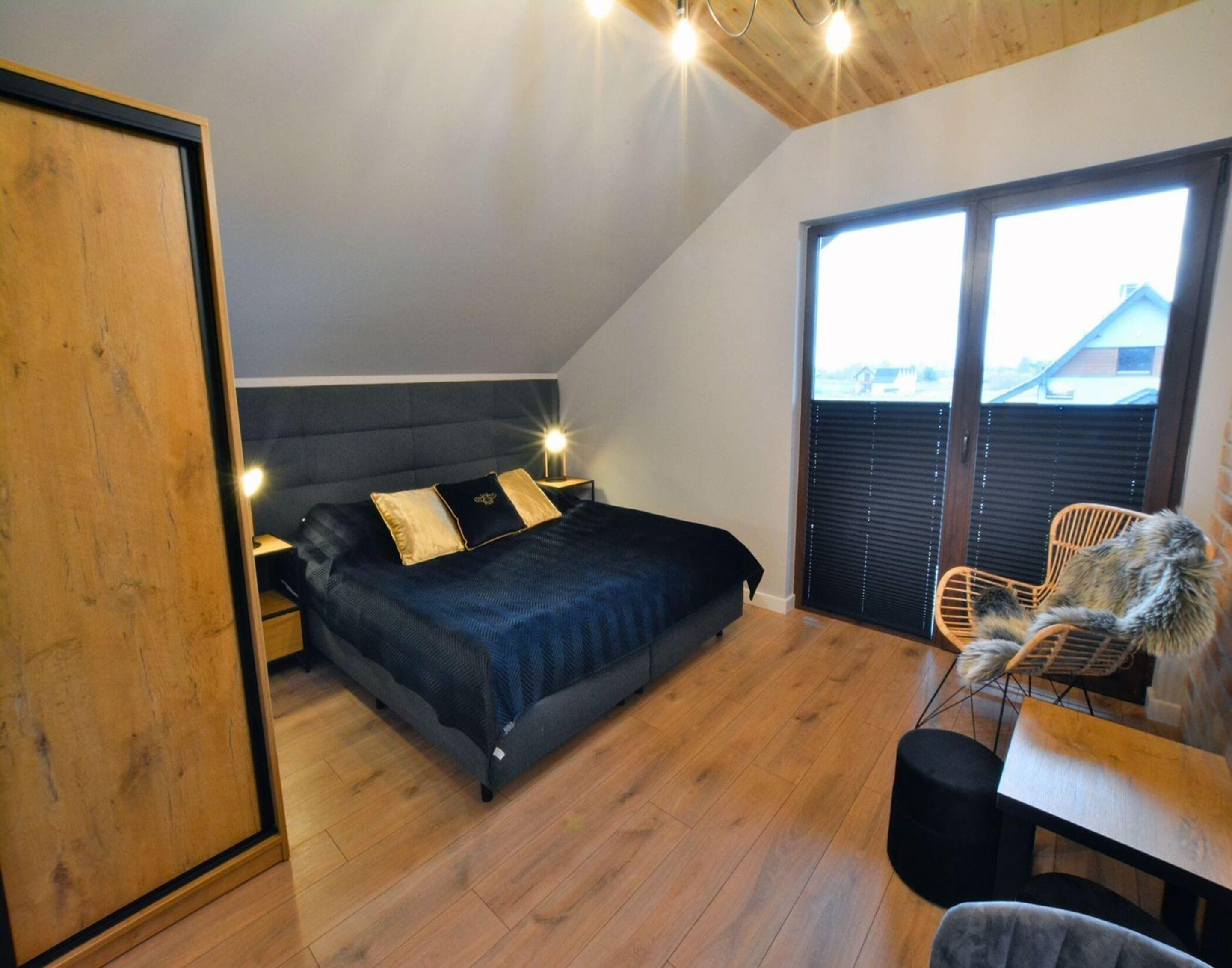 A bedroom with a bed, bedside tables, a wooden floor, a window with blinds, and a view of the outside.