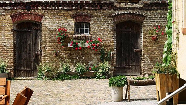 Un patio con suelo de ladrillo, bancos de madera y un edificio de ladrillo con ventanas arqueadas y macetas con flores.