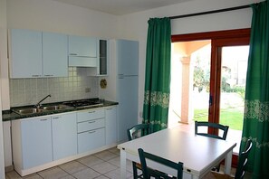 A compact kitchen with light blue cabinets, a sink, and a stove. There is a dining table with two chairs and a view of a garden through a doorway. - Flat With Pool in Badus Resort (Badesi)