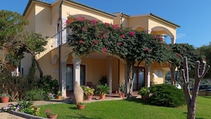 A two-story building with a balcony, surrounded by a well-maintained garden with various potted plants and flowers. - Flat With Communal Pool in the Residence Badus in Badus-formerly TUI Ferienhaus (Badesi)