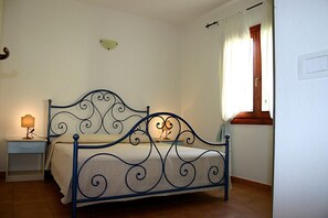A bedroom with a bed, a nightstand, a lamp, a window with curtains, and a wall switch. - Flat With Pool in Badus Resort (Badesi)