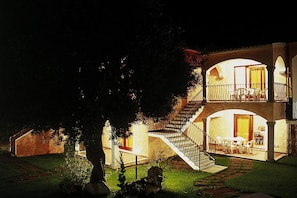 A two-story building with a staircase leading to an upper balcony, a dining area with tables and chairs, and a well-lit entrance. - Flat With Pool in Badus Resort (Badesi)