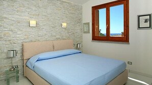 Un dormitorio con pared de piedra, una cama con colcha azul, ventana de madera y un cuadro en la pared.