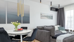 A modern living room with a grey sofa, black chairs, a white table with a vase of flowers, and a painting of a beach scene. - Premium Apartment With Pool and Sauna (Dziwnów)
