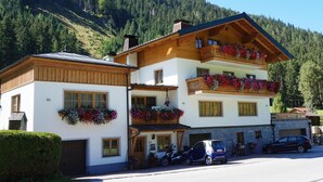 Ein zweistöckiges Gebäude mit hölzernen Balkonen, ein geparktes Auto davor und eine Bergkulisse im Hintergrund.