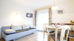 Ein modernes Wohnzimmer mit einer Couch, einem Esstisch, Stühlen, einem Fernseher und einem Fenster mit Vorhängen.