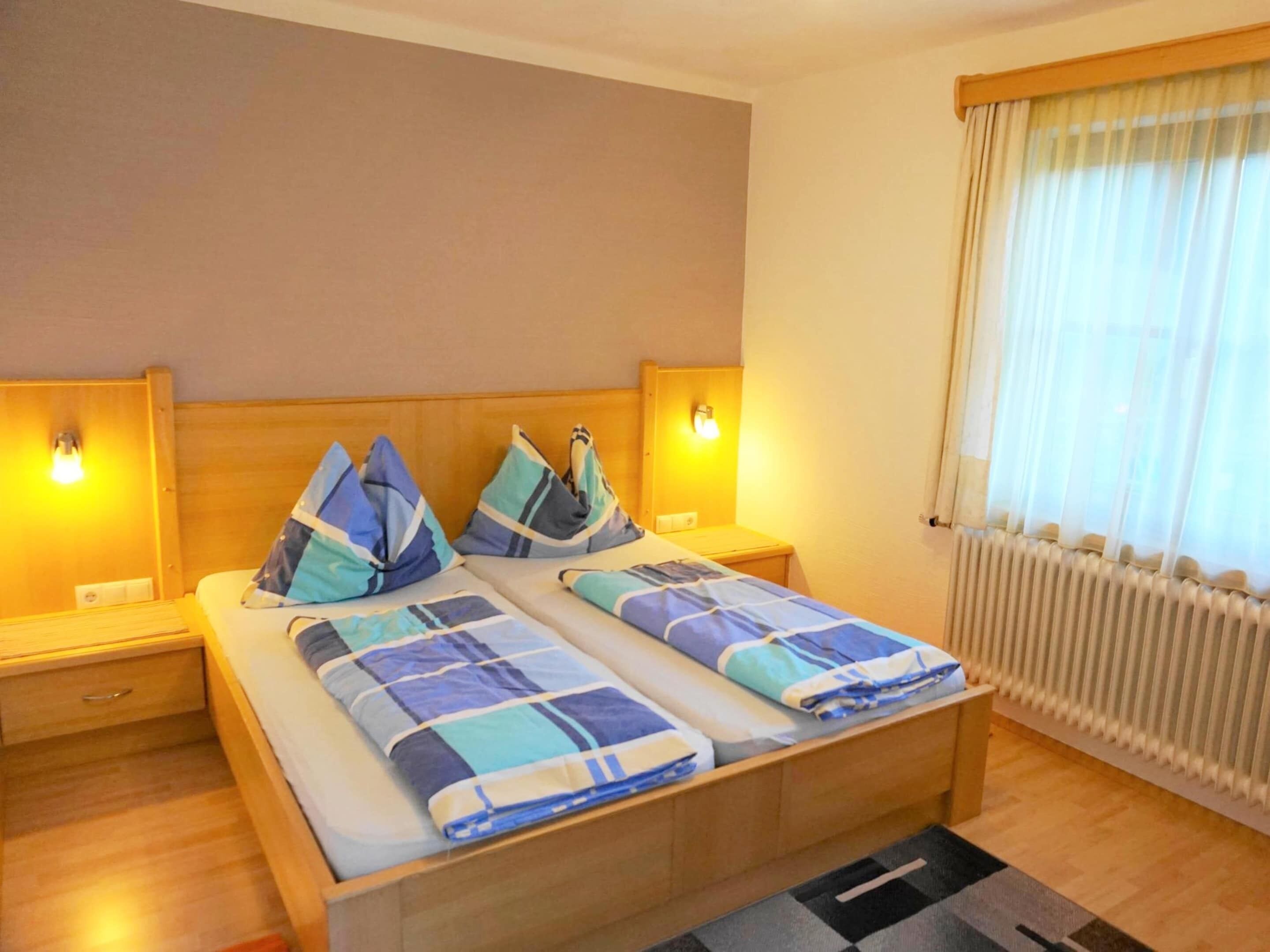 A double bed with a plaid bedspread, two bedside tables with lamps, a window with sheer curtains, and a radiator.
