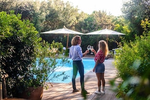 Twee personen staan bij een zwembad, met parasols en groen op de achtergrond.