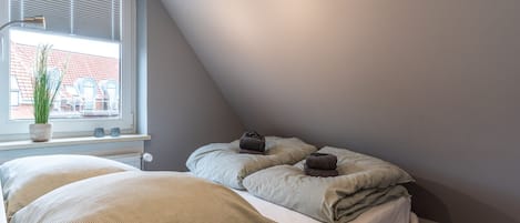 A bedroom with a bed, a window with blinds, a potted plant, and a bedside table with a lamp.