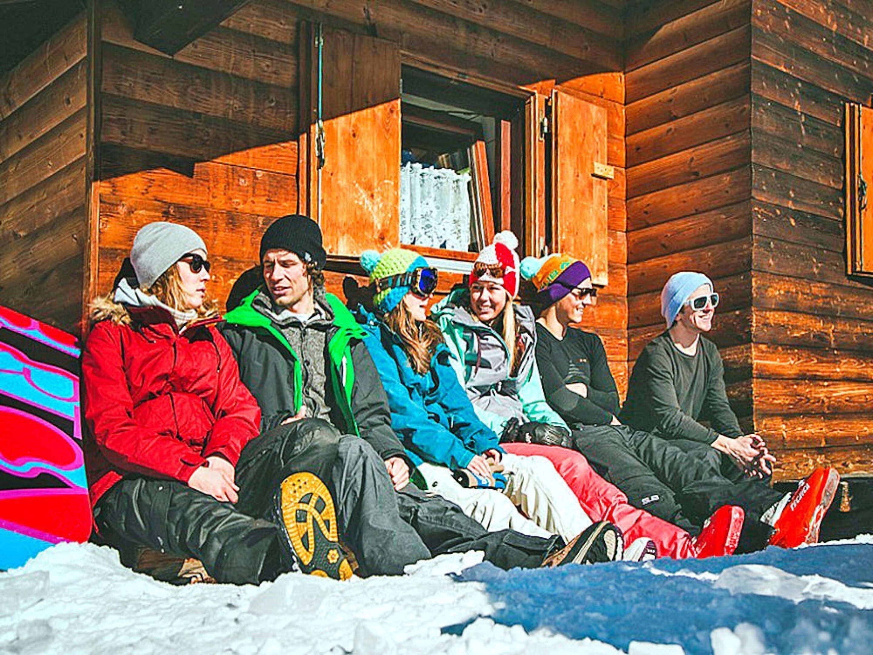 A group of people sitting on a wooden porch with a wooden cabin in the background.