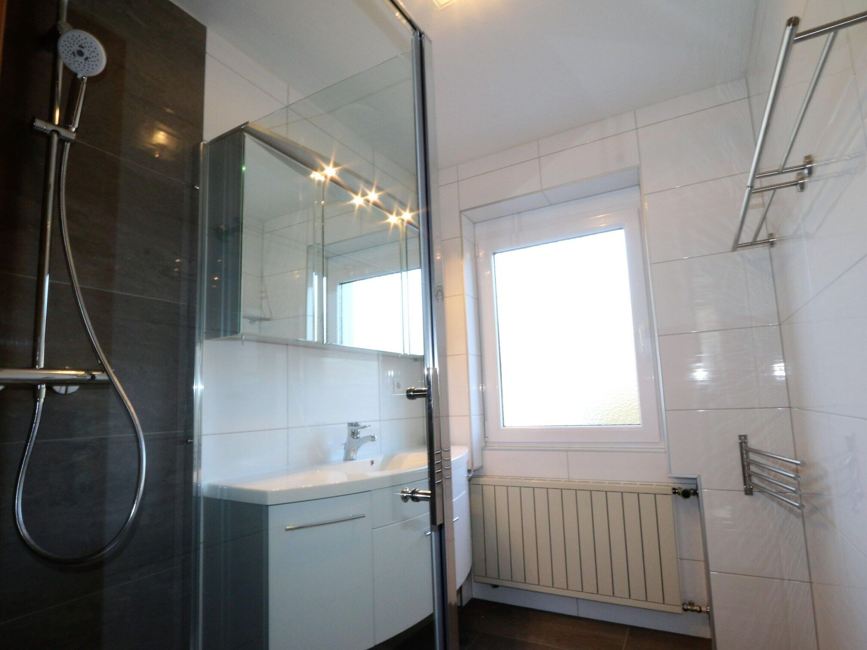 A modern bathroom with a glass shower enclosure, a large mirror, a window, and a white tiled floor.