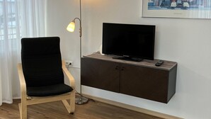 Una sala de estar moderna con un sofá negro, una lámpara de pie, un televisor de pantalla plana sobre un mueble de madera y un cuadro con la imagen de un restaurante en la pared.