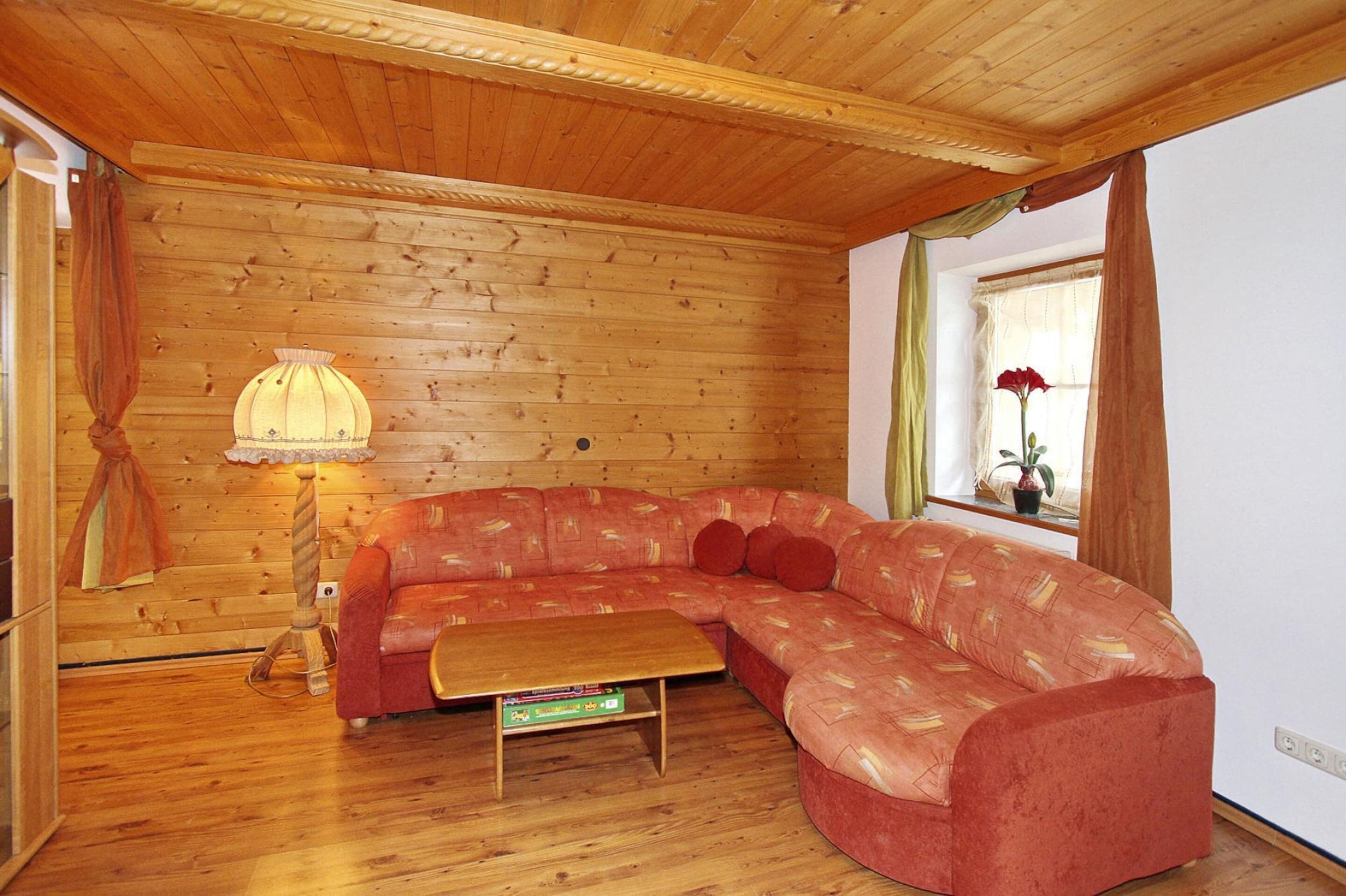 A cozy room with a wooden ceiling, a red sofa, a small table, a lamp, and a window with curtains.