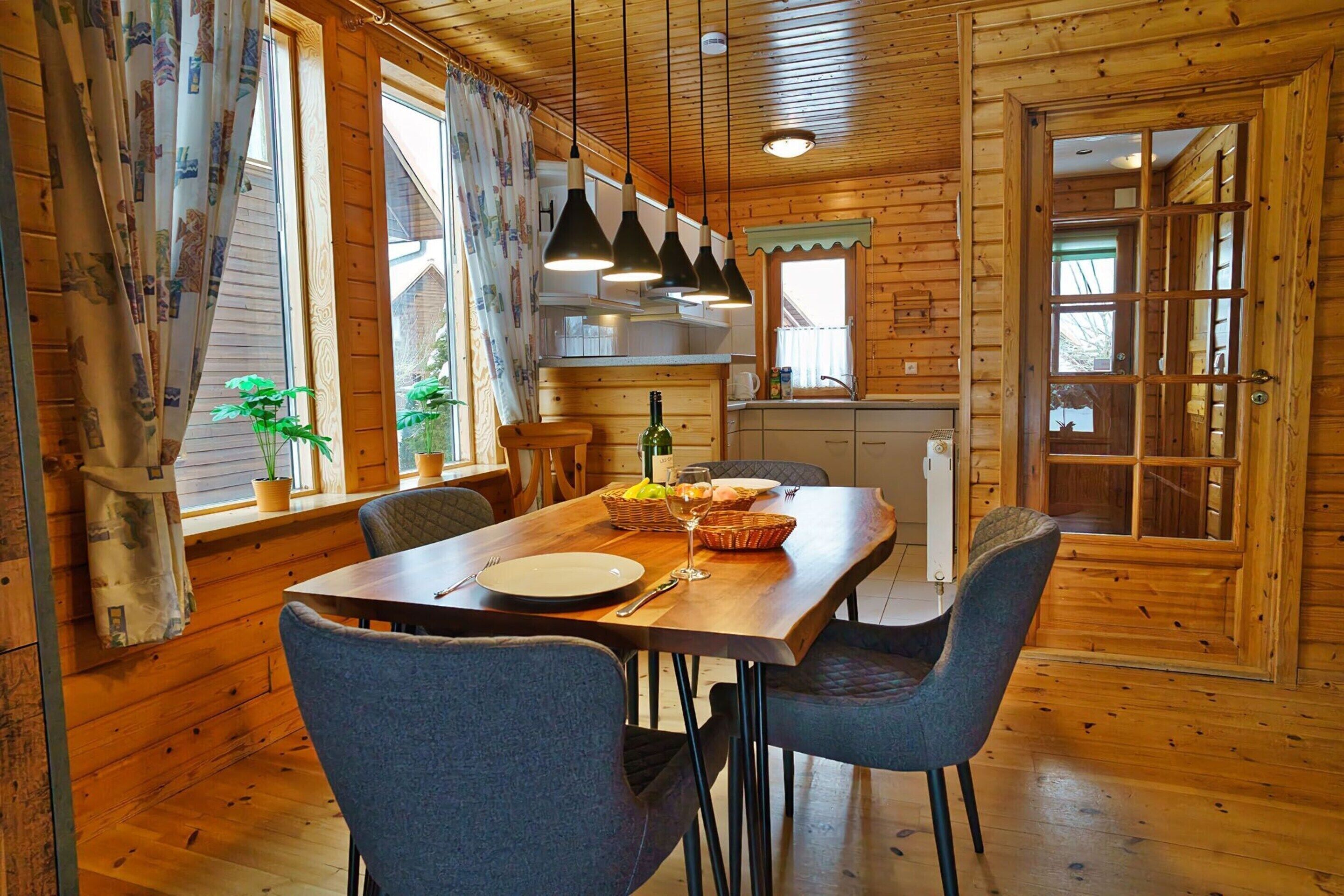 L’intérieur d’un chalet en bois comprend une table à manger pour quatre personnes, des chaises, un coin cuisine et une fenêtre avec des rideaux.