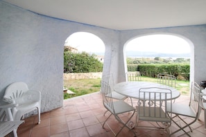 Eine Terrasse mit einem runden Tisch und Stühlen, einem weißen Kunststoffstuhl sowie Blick auf Grünflächen und entfernte Gebäude.