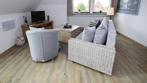 Un appartement moderne de style « loft » avec un plafond en pente, un coin salon confortable avec un canapé gris et un pouf en osier, un bureau en bois avec un téléviseur à écran plat et une grande fenêtre avec des rideaux.
