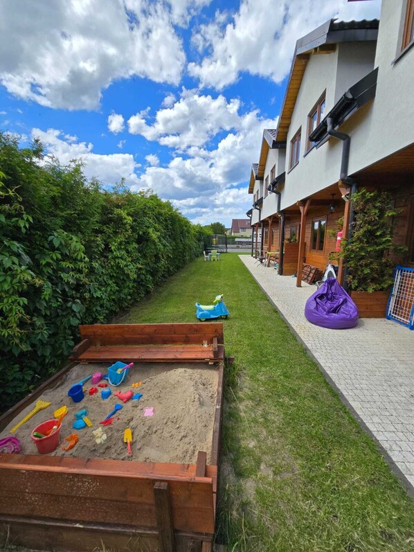 Una zona residenziale con un’area giochi in legno, un vialetto lastricato e una fila di case con facciate e balconi in legno.
