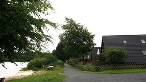A gravel path leading to a building with a dark roof and windows, surrounded by greenery and a body of water. - Ferienhaus mit Terrasse, Haustierfrei (Clausthal-Zellerfeld)