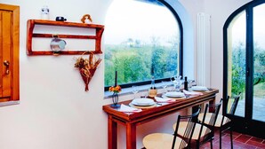 Un comedor con una mesa de madera dispuesta para cuatro personas, una ventana con vistas a follaje y una estantería con utensilios de cocina.