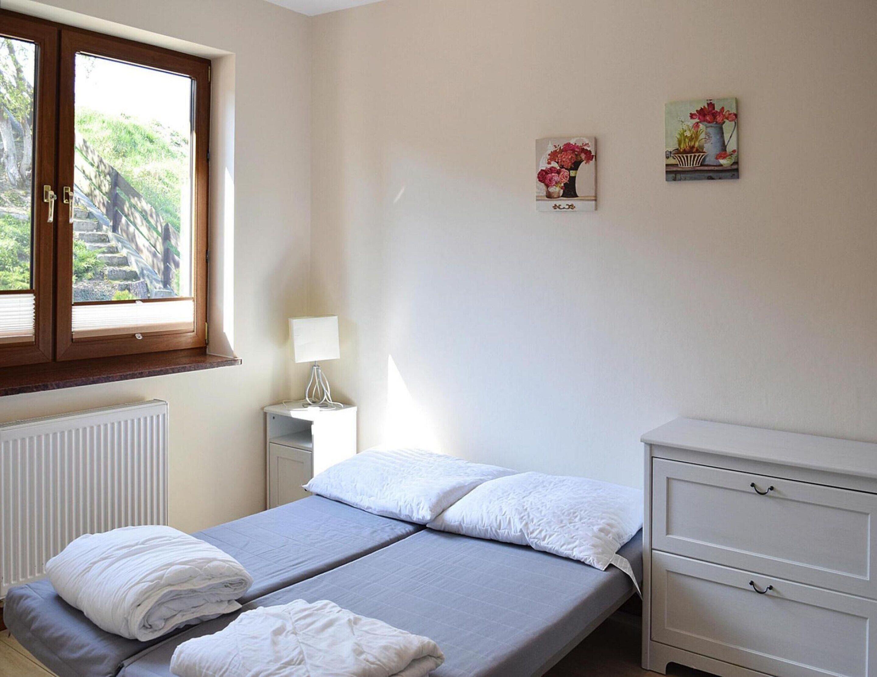 Une chambre avec deux lits, une fenêtre donnant sur un espace verdoyant, un radiateur, une table de chevet avec une lampe et une commode.