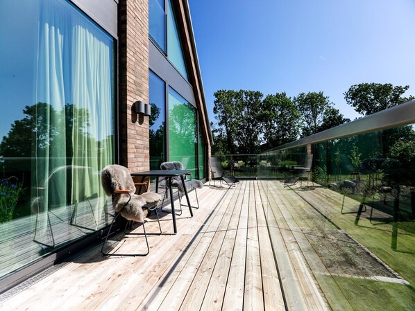 A wooden deck with outdoor furniture, large windows, and a view of trees. - Exklusives Landhaus auf Fehmarn (Fehmarn)