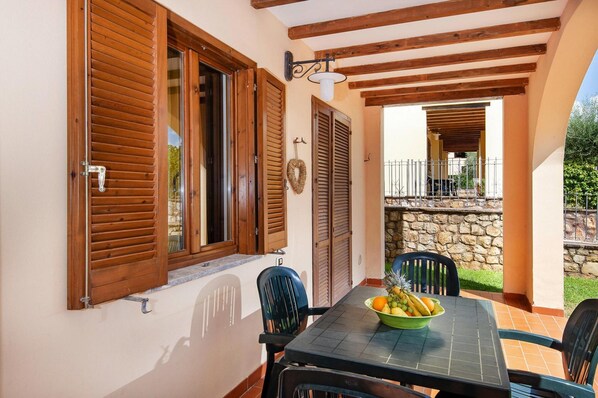 A dining area with a table set for two, a bowl of fruit, and a view of a stone wall and greenery.