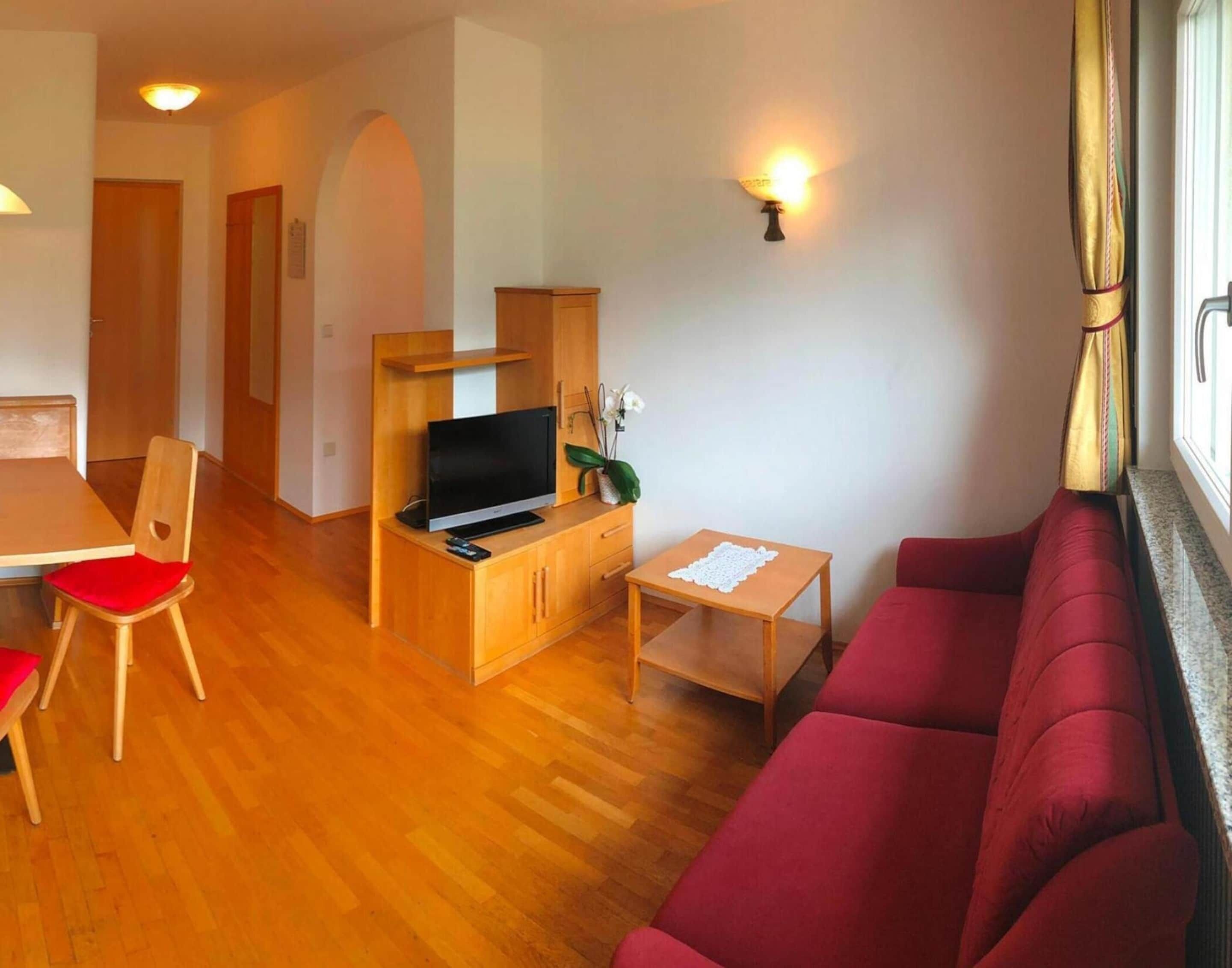 A compact living space with a wooden TV stand, a red sofa, a small table, and a dining area with a chair.