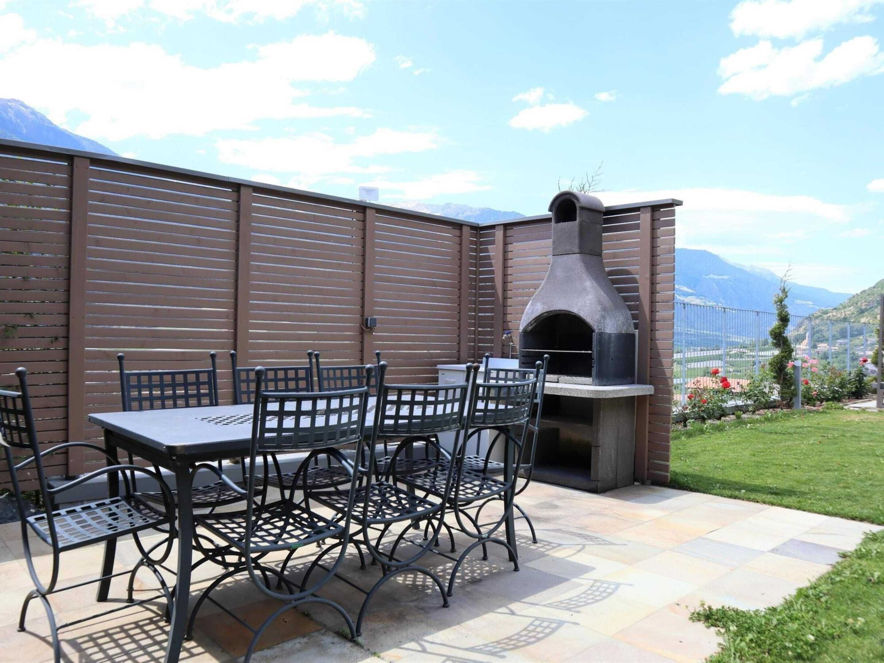An outdoor patio with a stone table and metal chairs, a stone oven, and a view of mountains in the background.