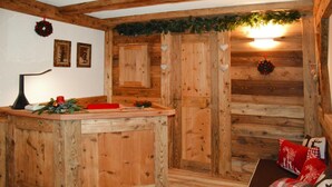 A rustic cabin interior with a wooden bar area, a couch with festive pillows, and a wall adorned with framed pictures and a wreath. - Apartment in Pellizzano With Paid Sauna (Pellizzano)