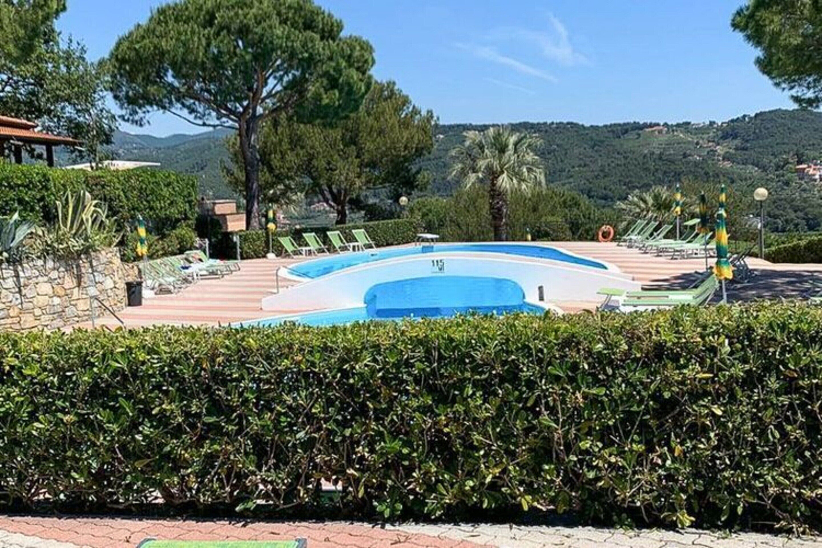 A swimming pool with lounge chairs, surrounded by hedges and trees, with a view of hills in the background.