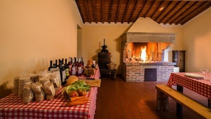 Een rustiek interieur met een bakoven, houten balken en een tafel met een geruit tafelkleed, eten en wijnflessen.