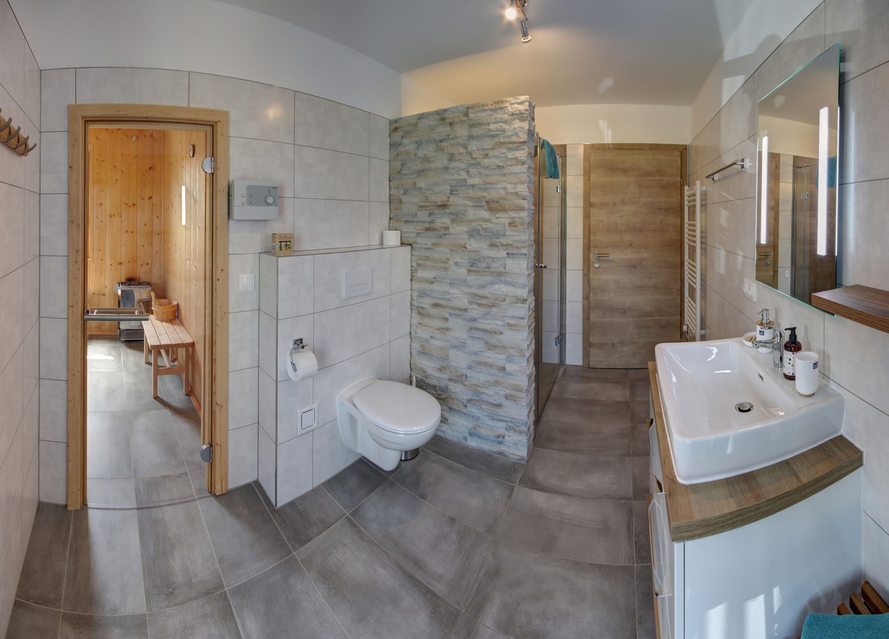 A modern bathroom with a stone accent wall, a wooden door, a toilet, a sink, and a shower area.