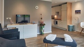 A modern living room with a flat-screen TV, a white entertainment unit, a large vase with flowers, and a bench with towels. - Das Ferienhus in Tonning (Toenning)