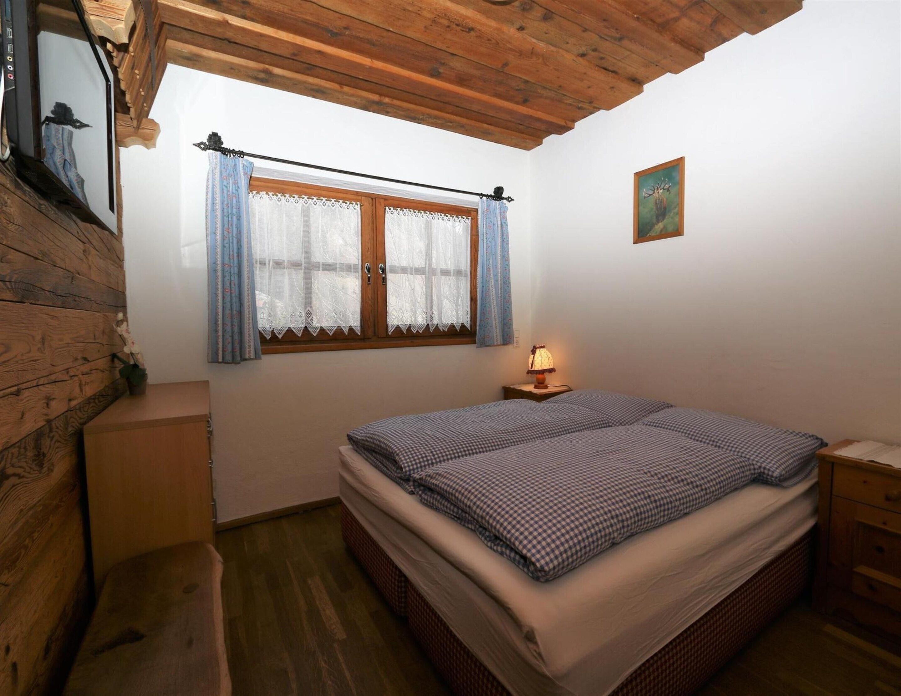 A cozy bedroom with a wooden ceiling, a double bed, a nightstand with a lamp, and a window with lace curtains.